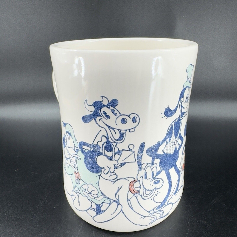 May include: White ceramic mug with a blue and white cartoon design featuring a cow, a dog, and a duck. The characters are in a playful pose and the mug has a rounded shape.