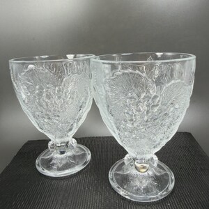 May include: Two clear, stemmed glasses with a textured grape and leaf design. The glasses have a heart-shaped bowl and a round base. The stem has a small, decorative bulb. The glasses are made of clear glass.