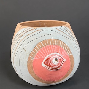 May include: A rounded ceramic planter with a wide opening, featuring a light tan base color. The exterior is decorated with thin, dark, horizontal lines and a prominent circular pink patch containing a raised, detailed eye illustration.