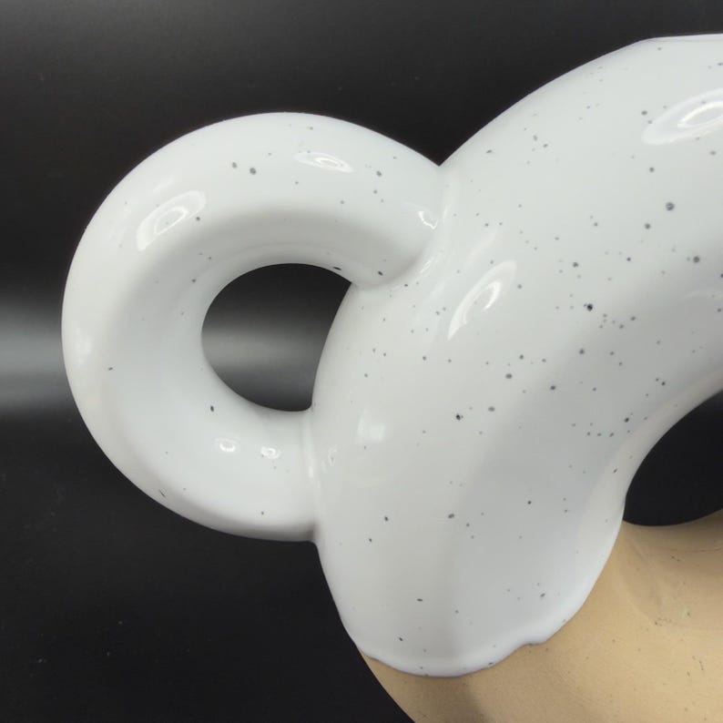 Peut inclure: Gros plan sur une anse en c&eacute;ramique blanche avec un design circulaire. La poign&eacute;e a une finition brillante avec de petites mouchetures noires. La poign&eacute;e est fix&eacute;e &agrave; une base en c&eacute;ramique beige, avec un fond sombre.