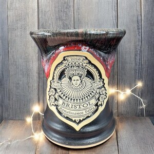May include: A ceramic mug with a flared top, featuring a black base and a red and black rim. The mug has a cream-colored emblem with a portrait, the words "Peace" and "Love", and "Bristol Renaissance Faire 2016".