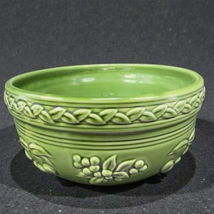 May include: A green ceramic bowl with a glossy finish. The bowl features a decorative leaf and floral pattern around the rim and lower portion. Horizontal lines and embossed details add texture to the design.
