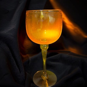 May include: A clear glass wine goblet with a yellow-orange hue, filled with a golden liquid. The goblet is on a black surface.