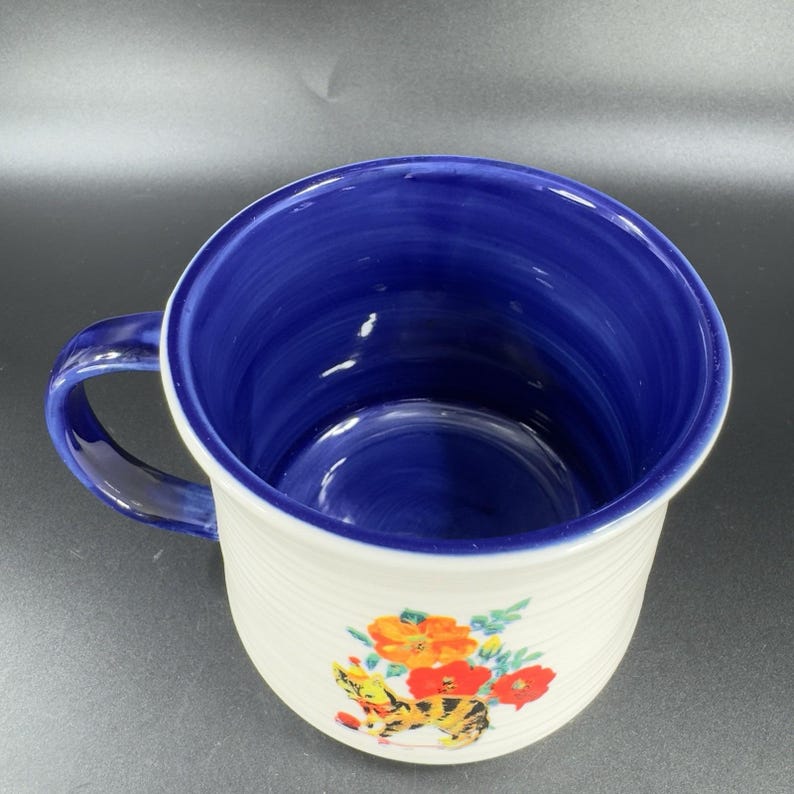 Puede incluir: Taza de cer&aacute;mica blanca con interior y asa azules. La taza presenta un dise&ntilde;o floral y de gato en naranja, rojo y amarillo. La taza tiene una textura acanalada en el exterior.