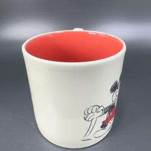 May include: Cream-colored ceramic mug with a red interior. The mug features a black and red sketch of Mickey Mouse on the exterior. The handle is small and white. The mug is a classic Disney collectible.