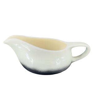 May include: A white ceramic gravy boat with a black rim. The boat has a rounded shape with a handle on the right side.