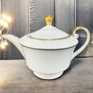 May include: White porcelain teapot with gold accents on the handle, spout, and lid. The lid is topped with a gold finial. A black and gold band encircles the top of the teapot. The teapot sits on a wooden surface.