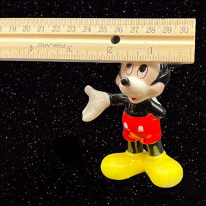 Puede incluir: Una figurita de cer&aacute;mica de Mickey Mouse, de pie con el brazo derecho extendido y la mano izquierda en la cadera. Lleva sus caracter&iacute;sticos pantalones cortos rojos con lunares blancos, zapatos negros y guantes blancos. La figurita mide aproximadamente 10 cm de alto.