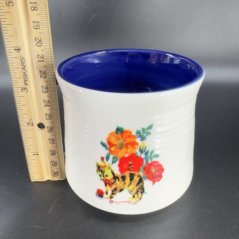 Puede incluir: Porta utensilios de cer&aacute;mica blanca con interior azul y un dise&ntilde;o caprichoso de gato y flores. El gato lleva un gorro de fiesta y est&aacute; rodeado de flores naranjas y rojas. El soporte mide unos 13 cm de alto.