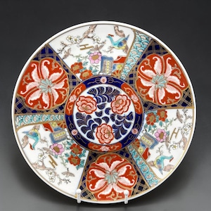 May include: A decorative white ceramic plate with a detailed floral and bird design. The plate features red, blue, and gold accents, with a central floral motif and surrounding panels with birds and flowers. The plate is supported by a white stand.
