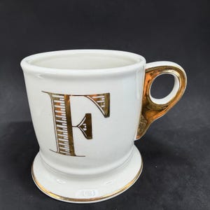 May include: White ceramic mug with a gold handle and the letter "F" in gold. The mug has a pedestal base and gold trim around the base and rim. The mug is on a black background.