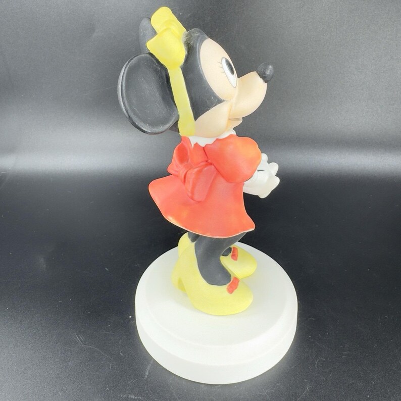 May include: A ceramic figurine of Minnie Mouse wearing a red dress with a yellow bow, yellow shoes, and a yellow bow in her hair. She is standing on a white base.