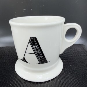 May include: White ceramic mug with a large black letter "A" on the front. The mug has a rounded base and a handle. The mug is a classic design, suitable for coffee or tea.
