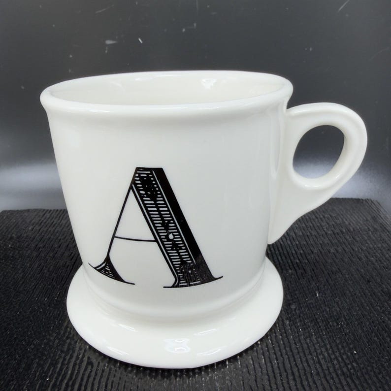 May include: White ceramic mug with a large black letter "A" on the front. The mug has a rounded base and a handle. The mug is a classic design, suitable for coffee or tea.