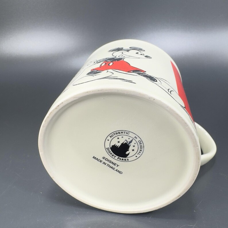 May include: White ceramic mug featuring a classic Mickey Mouse design in red, black, and white. The mug has a handle and a red stripe. The bottom of the mug has a Disney Parks logo and the text "Made in Thailand".