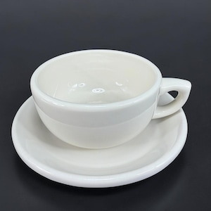 May include: A solid off-white ceramic coffee cup with a matching round saucer. The cup features a thick, rounded wall and a sturdy, angular handle on the side. The interior of the cup is smooth and deep, designed for holding hot beverages.