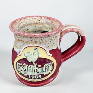 May include: A ceramic mug with a two-tone glaze featuring a deep red base and a cream-colored rim. The front displays a yellow oval emblem with a rooster illustration and the text Egg Harbor Cafe, Breakfast & Lunch, 1985, Geneva, Illinois.