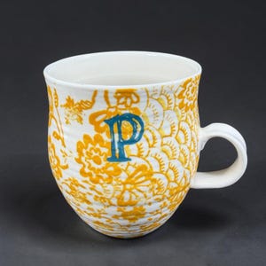 May include: A white ceramic mug featuring a ribbed texture and a yellow floral pattern covering the exterior. A prominent, bold, teal-colored letter P is centered on the front of the mug. The handle is white and extends from the right side.
