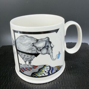 May include: White ceramic mug with a whimsical elephant illustration. The elephant is gray with a black harness and is holding a blue watering can. The mug has a black border and a colorful patterned base.