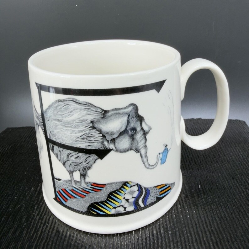 May include: White ceramic mug with a whimsical elephant illustration. The elephant is gray with a black harness and is holding a blue watering can. The mug has a black border and a colorful patterned base.