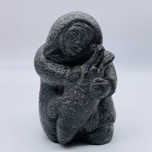 May include: A dark gray stone sculpture of a person in a hooded parka embracing a smaller figure. The sculpture has a textured surface and is set against a white background. The figures are depicted with simple facial features.