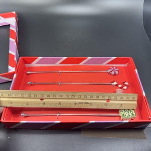 May include: A set of three cocktail stirrers with decorative candy-themed tops, presented in a red and pink striped box. The stirrers are approximately 10 inches long. The box is red with pink and white diagonal stripes.