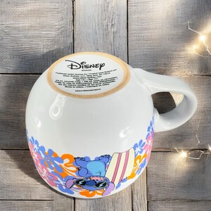 May include: A white ceramic mug with a blue and orange floral design. The mug features a cartoon image of Stitch from Lilo & Stitch, wearing sunglasses and holding a surfboard. The bottom of the mug has the Disney logo and product information. "Produced under license by/ Produit sous licence par Silver Buffalo, LLC, New York, NY 10010 www.silverbuffalo.com Age 14+/ Age 14+ 24 oz. ceramic mug / Tasse en c&eacute;ramique 24 oz. Safe to use in microwave and dishwasher.  Not for use in oven or on stovetop.  Va au micro-ondes et au lave-vaisselle.  Ne pas utiliser au four ou sur la cuisini&egrave;re. Made in China / Fabriqu&eacute; en Chine. CCL1140333072019"