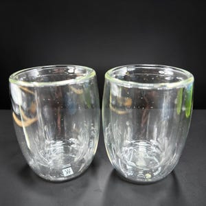 May include: Two clear, double-walled glass tumblers with a rounded base and a smooth rim. Each glass features a small, square white logo with a twin-figure icon near the bottom base, indicating a double-walled construction for thermal insulation.