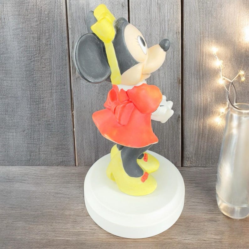 May include: A figurine of Minnie Mouse wearing a red dress with a yellow bow and yellow shoes. The figurine is standing on a white base.