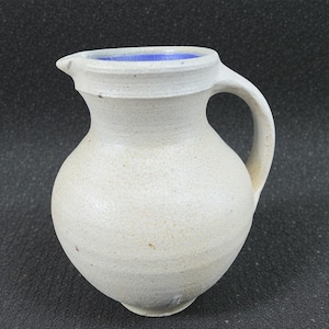 May include: A handmade, off-white ceramic pitcher with a rounded body and a handle. The pitcher has a spout and a blue interior. The exterior has a speckled texture. The pitcher is suitable for serving beverages.