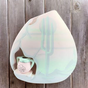May include: A white ceramic vase shaped like a mountain with a cutout for a smaller vase. The larger vase has a painted desert scene with a green cactus and a white moon. The smaller vase is light pink with green and blue accents and a face.