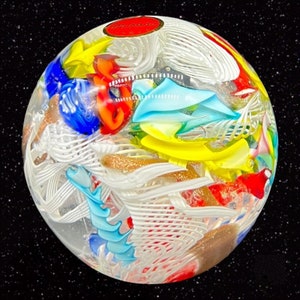 May include: A colorful glass paperweight with a white, red, blue, yellow, and gold design. The paperweight is round and has a clear glass exterior. The text "Murano" is visible on the paperweight.