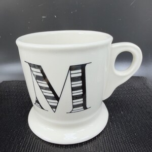 May include: White ceramic mug with a black letter "M" design. The mug has a rounded base and a handle. The letter "M" is large and stylized with black and white stripes. The mug is suitable for coffee, tea, or other beverages.