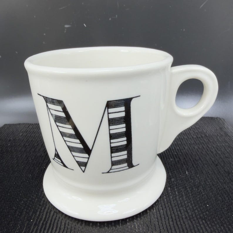 May include: White ceramic mug with a black letter "M" design. The mug has a rounded base and a handle. The letter "M" is large and stylized with black and white stripes. The mug is suitable for coffee, tea, or other beverages.