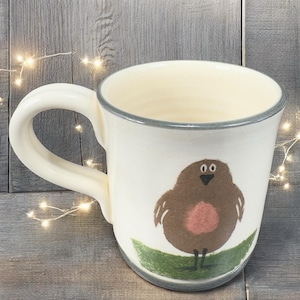 May include: White ceramic mug with a hand-painted robin design. The mug features a brown bird with a red breast, perched on a green leaf. The rim and handle are a light cream color, with a gray trim. The mug is approximately 4 inches tall.