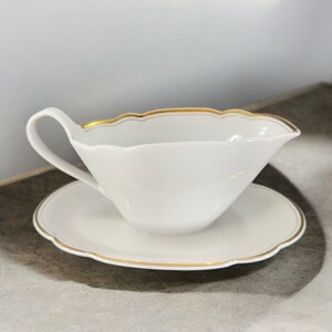 May include: A white porcelain gravy boat with a gold trim and a matching saucer. The boat has a scalloped edge and a handle.