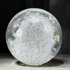 May include: A clear glass sphere with internal white cloud-like formations and numerous air bubbles. The sphere is illuminated, creating highlights and refractions. The overall effect is a decorative object with an ethereal quality.