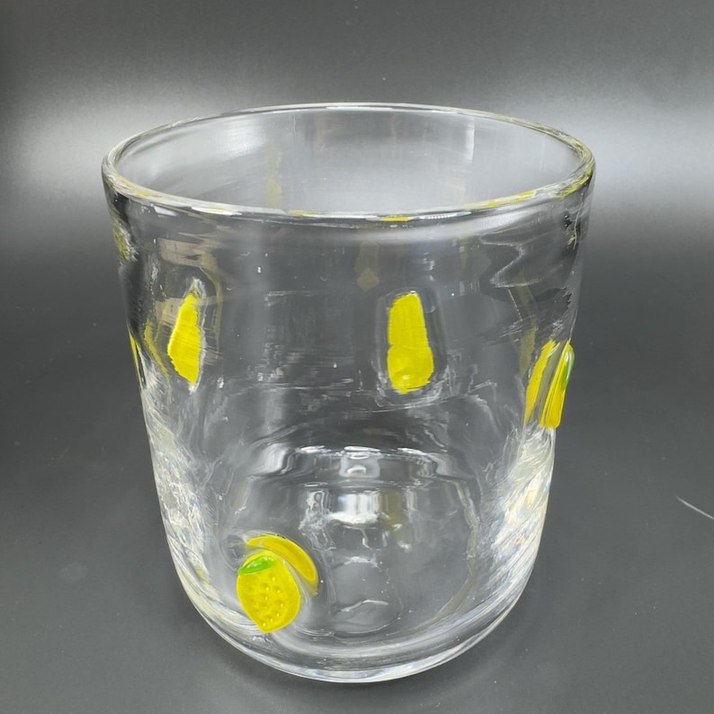 May include: A clear glass tumbler with yellow lemon-shaped designs. The glass has a slightly rounded shape and is decorated with several yellow lemon shapes. The glass is empty and sits against a dark background.