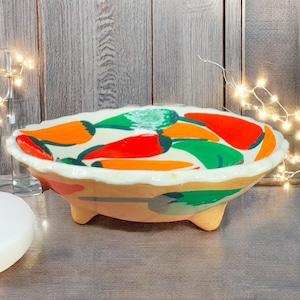 May include: A ceramic serving bowl with a scalloped rim and three small feet. The bowl is painted with a pattern of red, orange, and green chili peppers on a white background. The rim is white, and the exterior has tan and gray accents.