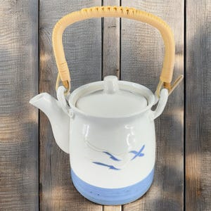 May include: White ceramic teapot with a blue and white bird design. The teapot has a woven bamboo handle and a lid.