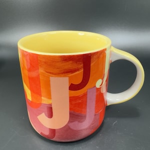 May include: A ceramic mug featuring a repeating pattern of the letter J in various shades of red, orange, and pink. The mug has a pale yellow interior and a white handle, with a smooth, glossy finish and a cylindrical shape.