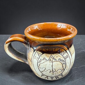 May include: A ceramic mug with a glossy brown glazed rim and handle, transitioning to a cream-colored base. A circular emblem on the front features a bear silhouette, mountain peaks, and the text ZAKOPANE TATRY embossed in the clay surface.