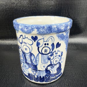 May include: A ceramic planter with a white base and a blue rim. The planter features a hand-painted design of cartoon pigs in blue. The pigs are wearing scarves and have hearts above their heads.
