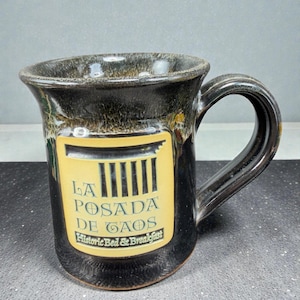May include: A black ceramic mug with a curved handle. The mug features a yellow rectangular label with the text "LA POSADA DE GAOS Historic Bed & Breakfast" in black. The mug has a dark brown, speckled glaze.