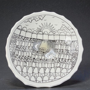 May include: White ceramic bowl with a scalloped edge, featuring a black and white illustration. The artwork depicts a sun, trees, birds, and a crowd of people. The bowl's design includes a village scene at the bottom.