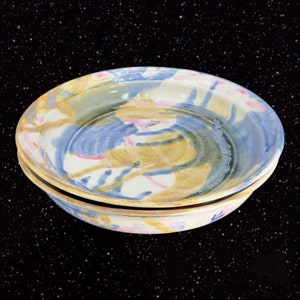 May include: Two ceramic pie plates with a hand-painted design in shades of blue, yellow, and pink. The plates are stacked on top of each other.