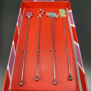 May include: A set of four cocktail stirrers with candy-themed toppers, including a swirl, a candy cane, a wrapped candy, and a green and white candy. The stirrers have long, slender silver stems and are presented in a red box.