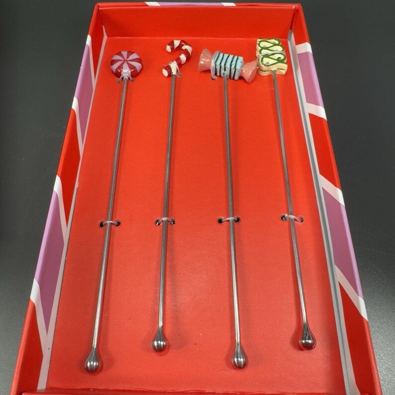 May include: A set of four cocktail stirrers with candy-themed toppers, including a swirl, a candy cane, a wrapped candy, and a green and white candy. The stirrers have long, slender silver stems and are presented in a red box.