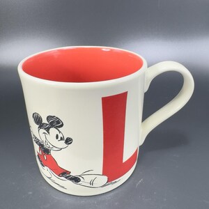 May include: Cream-colored ceramic mug with a red interior and a large red letter "L". The mug features a black and red illustration of Mickey Mouse. The handle is white.