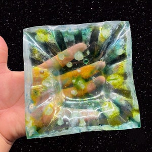 May include: A square, clear glass dish with a wavy, rippled surface. The dish has a light blue, green, and yellow swirl pattern with white and yellow flecks throughout.